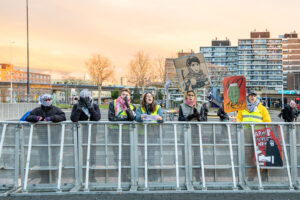 Demonstranten achter een hek, deels gemaskerd, met een paar borden met tekst.